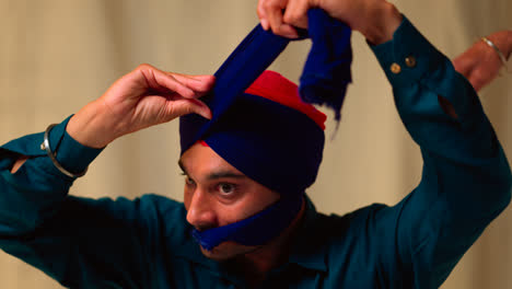 Fotografía-De-Estudio-De-Un-Hombre-Sikh-Mayor-Que-Ayuda-A-Un-Hombre-Sikh-Más-Joven-A-Atar-La-Tela-Para-Un-Turbante-Contra-Un-Fondo-Liso,-Filmada-En-Tiempo-Real-4